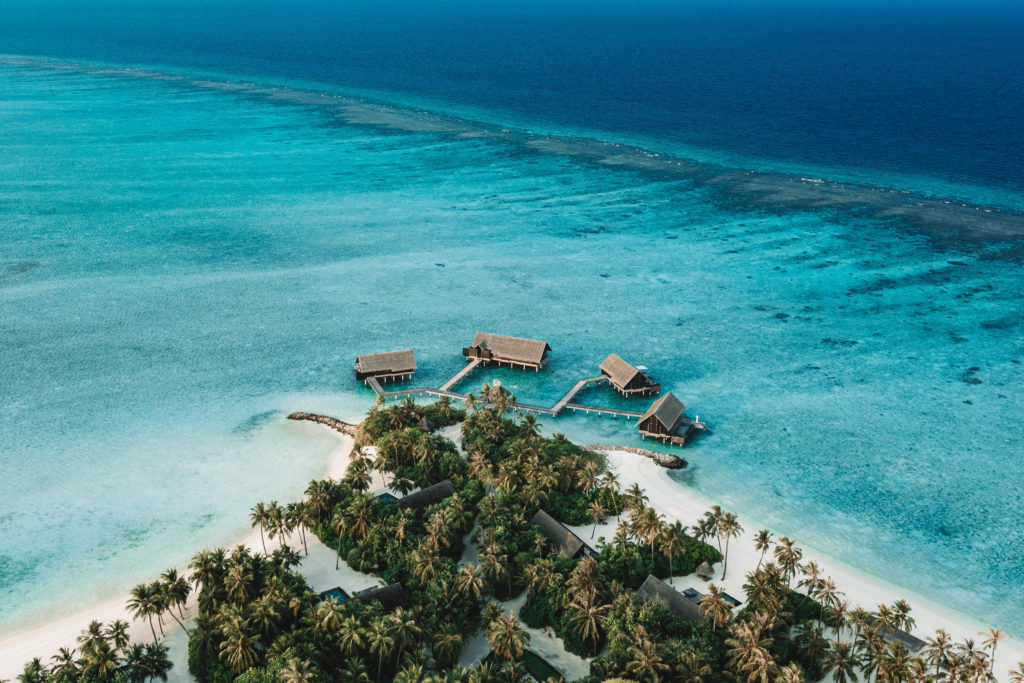 Aerial view of 4 overwater villas extending off the palm tree and white sand island.