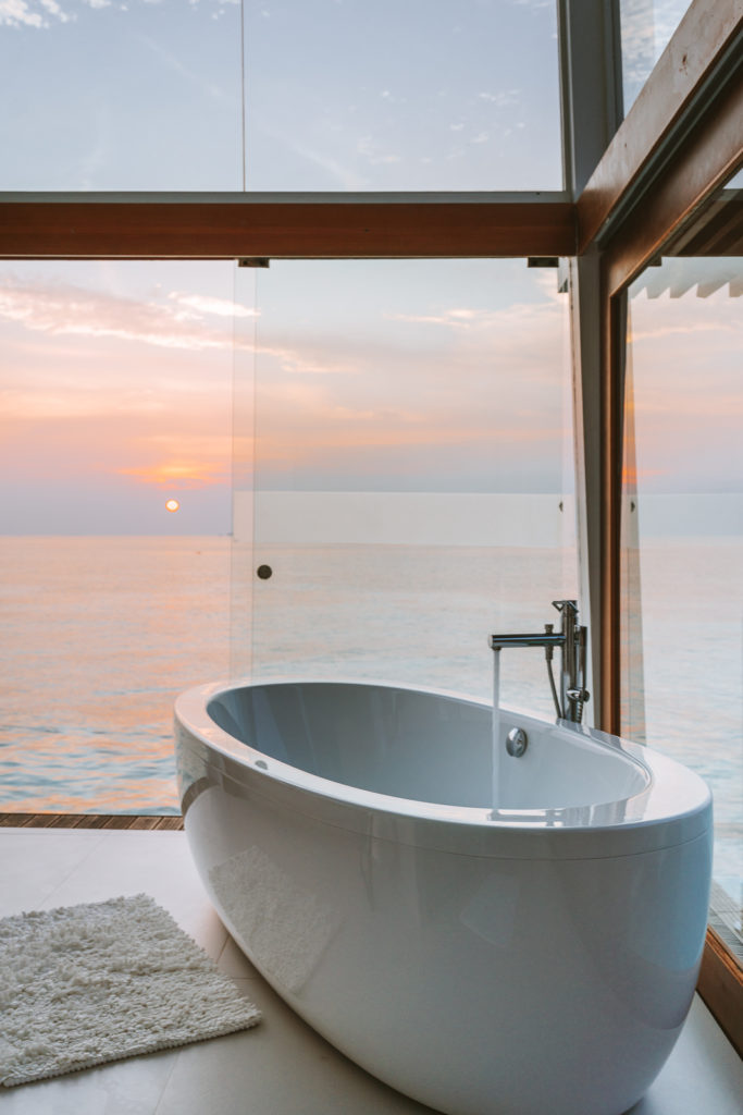 Bathtub running in a glass walled bathroom, with sunset views over the lagoon