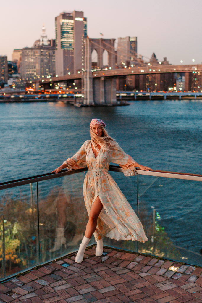 Sarah / SALTY LUXE stands in a flowy bohemian dress at sunset on a rooftop overlooking Brooklyn bridge & Manhattan skyline. 