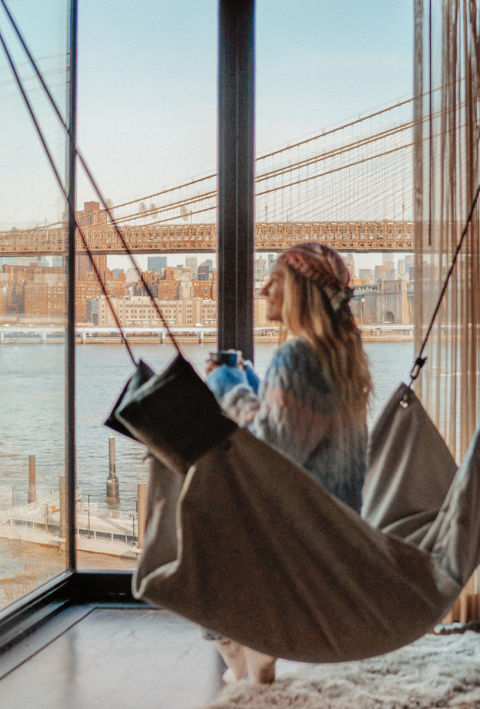 Sarah sitting in an indoor hammock looking over the river, sipping a coffee with the Brooklyn Bridge & Manhattan skyline in the background.