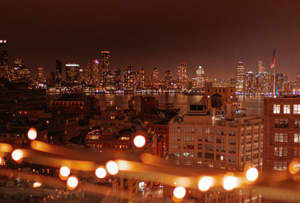 City skyline at night with fairylights twinkling from a balcony & office building lights shine in the background.