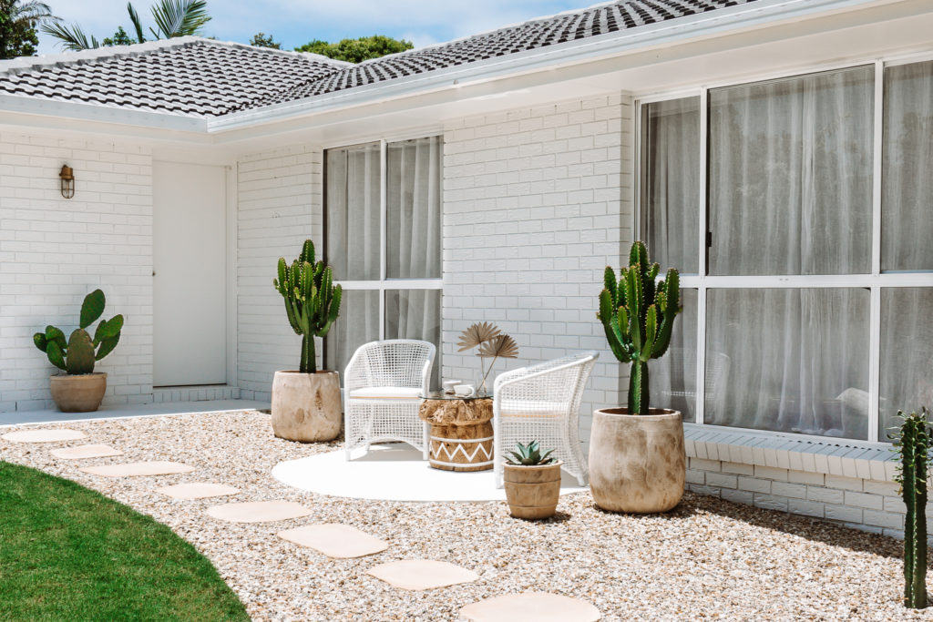 Rattan chair & large round natural tree trunk table with glass top, sit atop a hand circle cement terrace at the front of the house. Space sits among immersive gardens, with cacti surrounding & natural pavers.