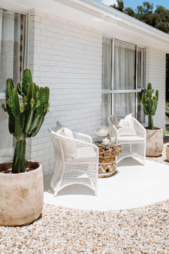 Rattan chair & large round natural tree trunk table with glass top, sit atop a hand circle cement terrace at the front of the house. Space sits among immersive gardens, with cacti surrounding & natural pavers