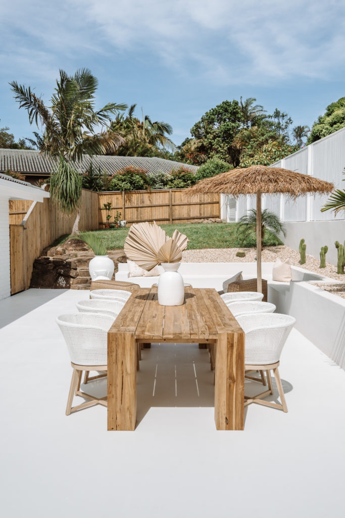 Large timber dining table sits sun drenched in the afternoon light, surrounded by cacti gardens. Tabletop has a large white vase & dried palms for texture.