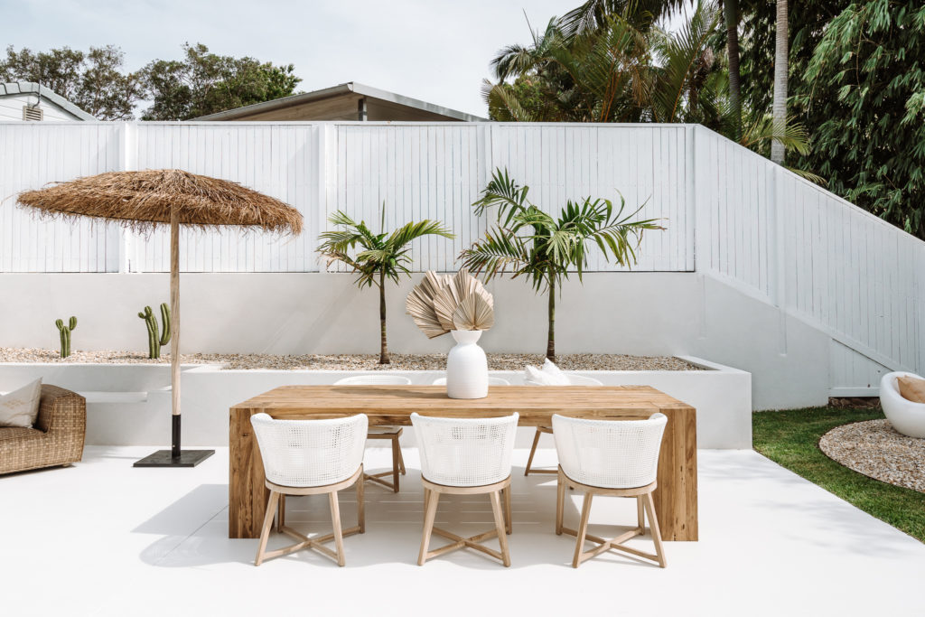 Outdoor block dining table with white rattan chairs framed by palm trees to the back, woven umbrella to the left & fire pit to the far right.