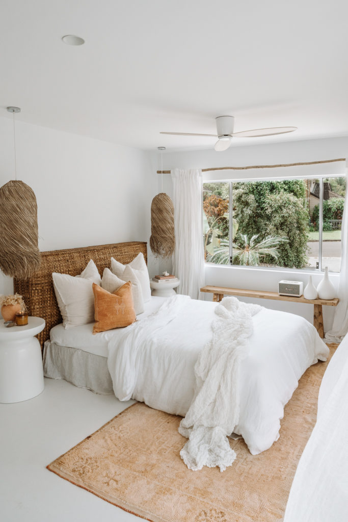 Queen bed with rattan bedhead, white linen & large moroccan rug in earthy tones dressing the underside of the bed. Natural hanging lights & vintage elm bench finish off the look.