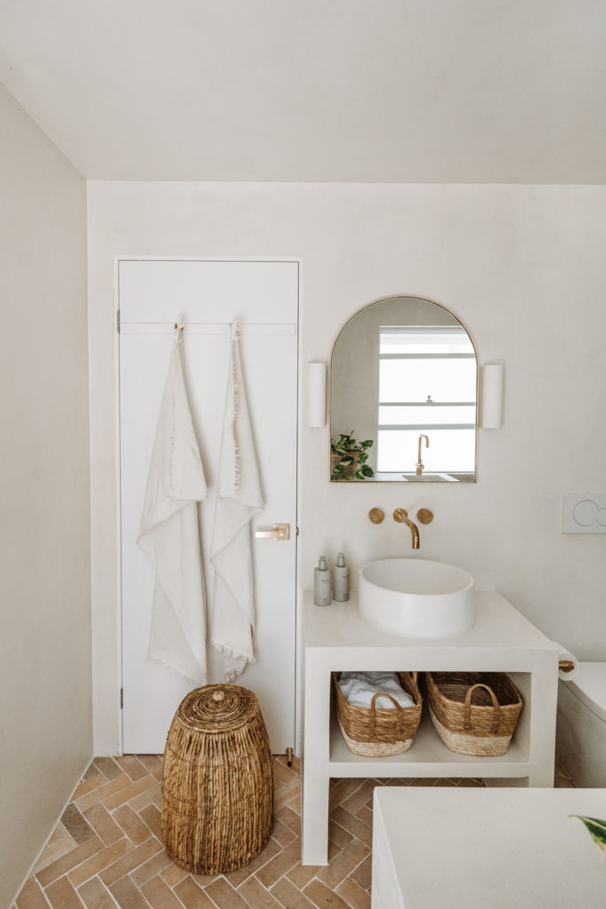 Freestanding rendered concrete basin & circular sink, with large arched brass rimmed mirror, rattan baskets & white linen hanging from the back of the door. 