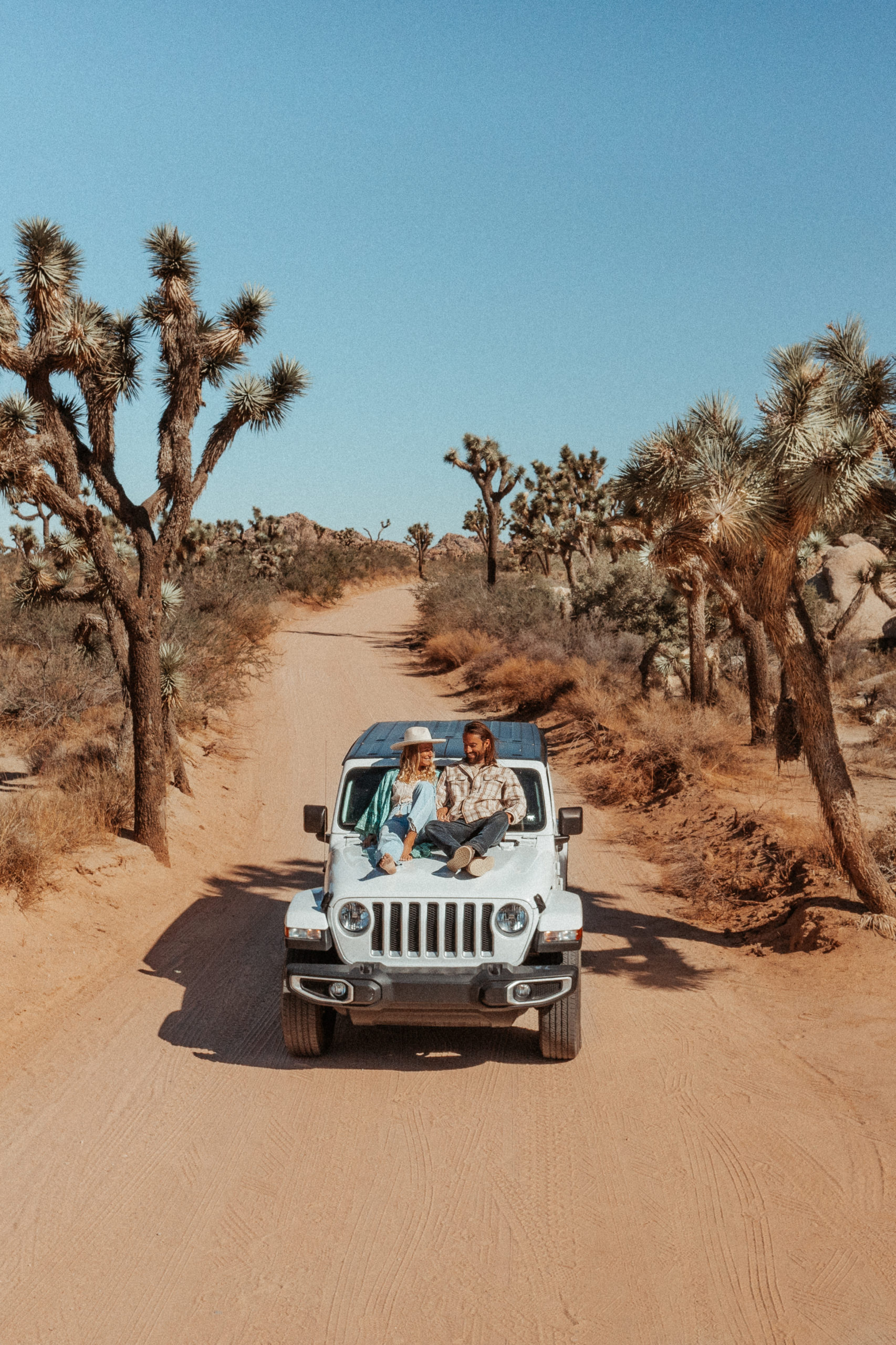 Sarah and Chesh on the front of a white Jeep, in Joshua Tree National Park