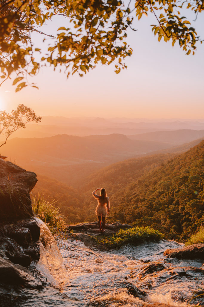 A woman in white overlooking a green valley from the top of a waterfall at sunset.