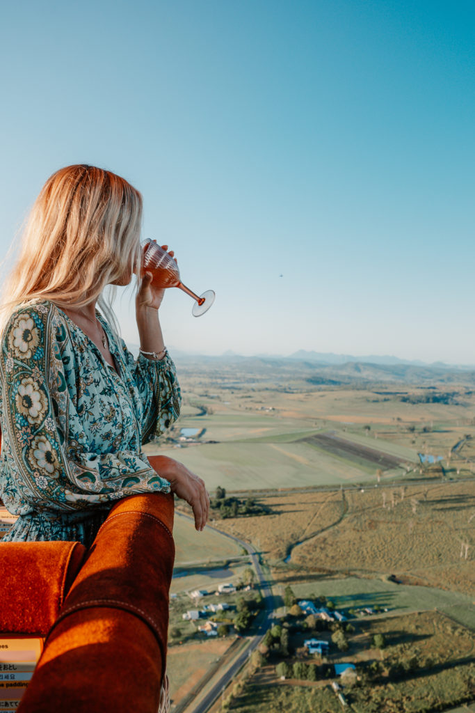 A woman in a green and white patterned shirt drinking Champagne with a view from hot air balloon looking out onto the green rolling countryside
