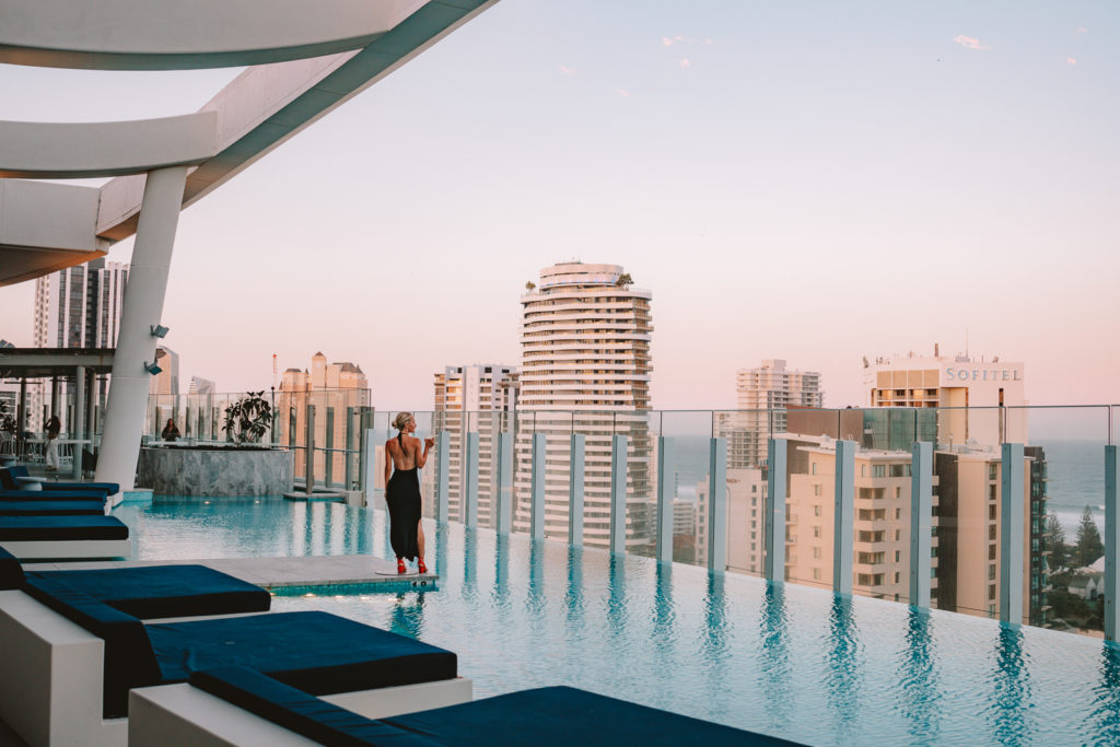 A woman in a black dress holding a glass of wine on the edge of a blue rooftop pool at The Darling Hotel. A city skyline is in the background