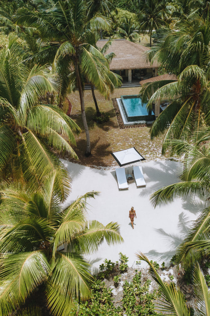 Aerial view of woman walking towards luxury villa and pool, surrounded by palm trees.