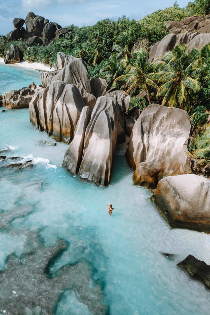 Aerial view off woman in water shallows with giant boulders and palm trees behind.
