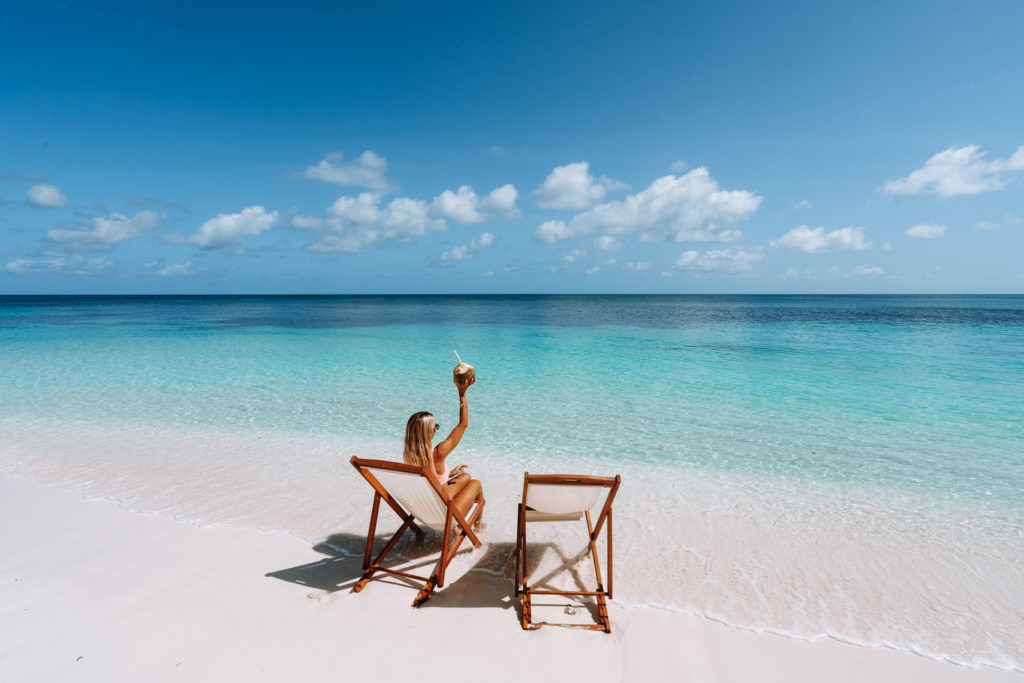 Woman in deck chair raising a coconut and facing the clear, calm, blue ocean.