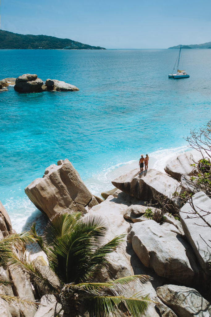 Couple standing on top of boulders, overlooking blue ocean and a sailing boat.