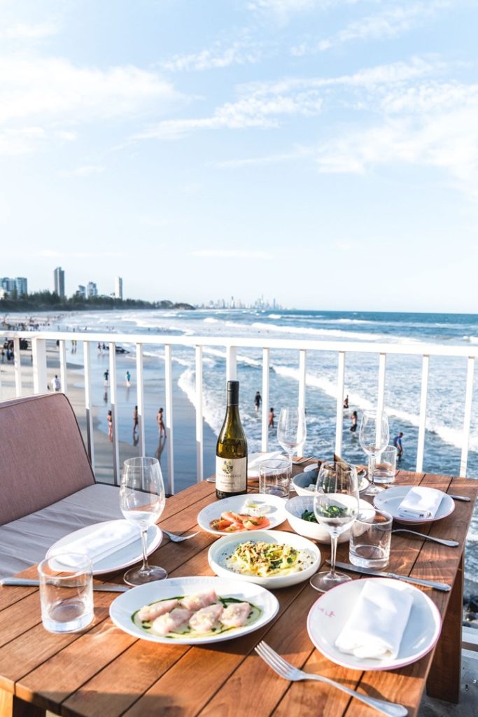 Many plates of food in white dishes on a wooden table overlooking a beach, at The Tropic restaurant 