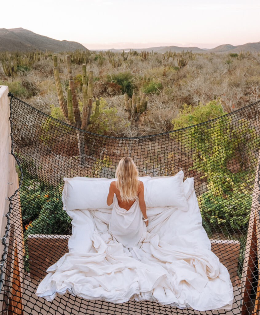 Woman in white bedsheets and a suspended hammock netting, overlooking cactus fields from her room at Paradero Hotel.