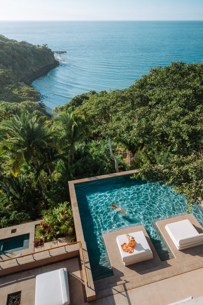 Woman sits by pool and man swims in pool overlooking the jungle and ocean.