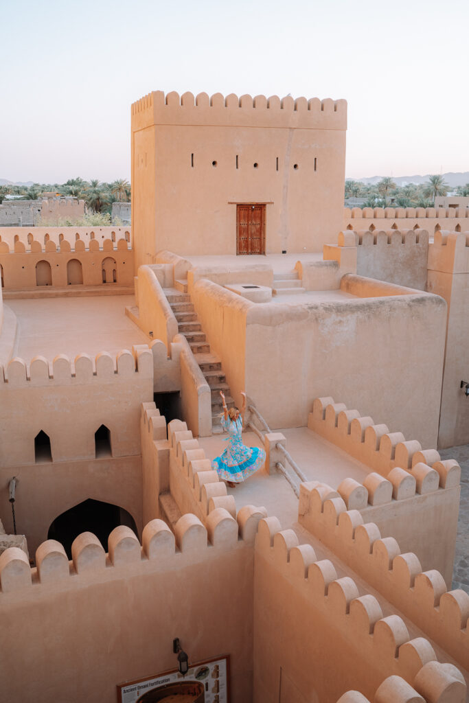 Woman twirls in a blue dress on top of a fortress that forms part of Nizwa Fort in Oman. The fort is orange terracotta coloured