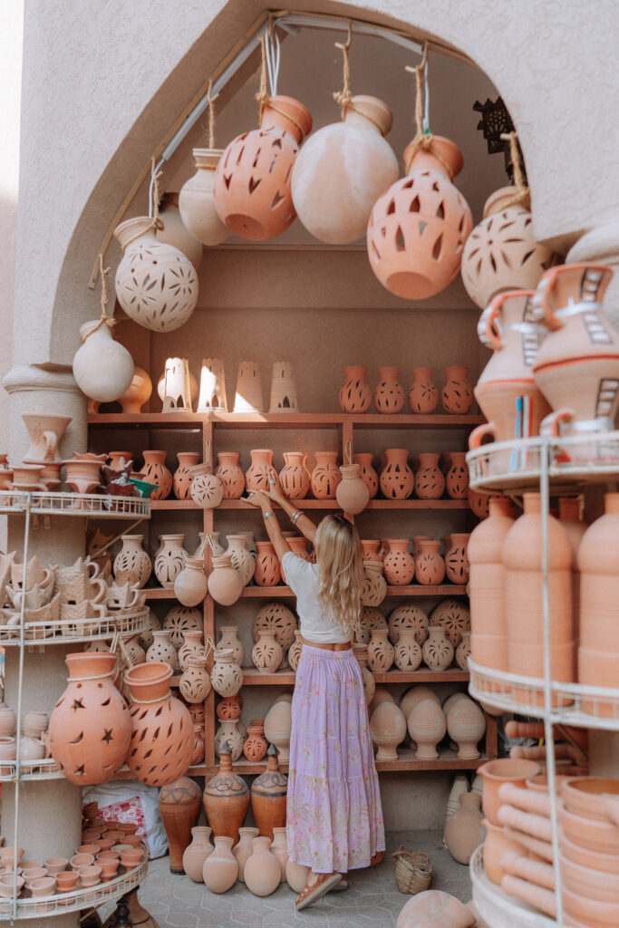 Woman in purple skirt reaching up and picking up a terracotta vase from a shelf inside the Nizwa Souks