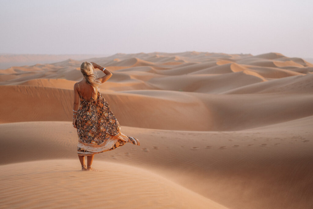 Woman stands on sand dunes looking over endless sand dunes in the Oman desert.m Wearing a floral print dress that's blowing in the wind