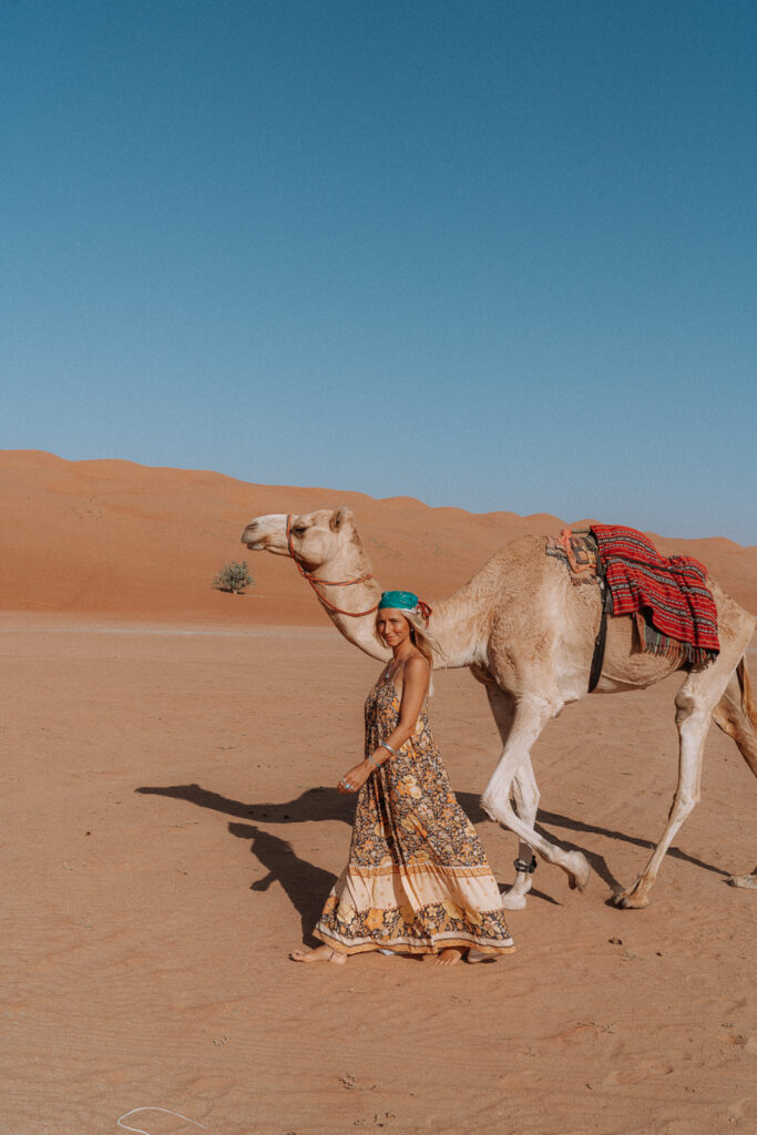 Woman in floral dress walks besides a camel in Wahiba Sands desert
