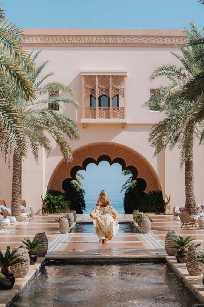 Woman walks away in a dress inside of an courtyard at Shangri-La Al Husn. A shallow pool in front and behind her with seating along the edge of the countyard. The sea is visible in the distance through a stone archway