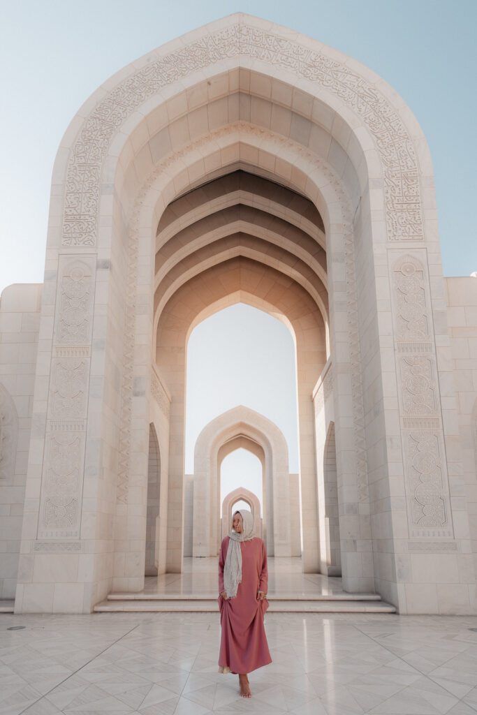 Woman in pink dress and a head scarf walks beneath large stone archways at the Grand Mosque
