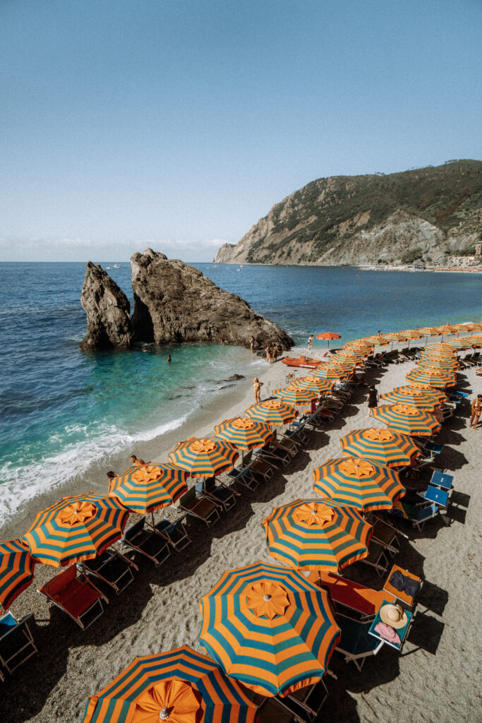 View of the beach in Monterosso. Large rock juts out into the sea. Umbrellas line the beach. People swim and sunbathe on the beach.