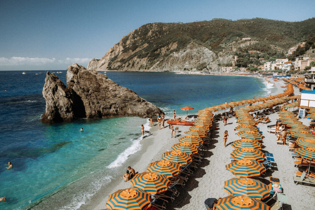 View of the beach in Monterosso. Large rock juts out into the sea. Umbrellas line the beach. People swim and sunbathe on the beach.