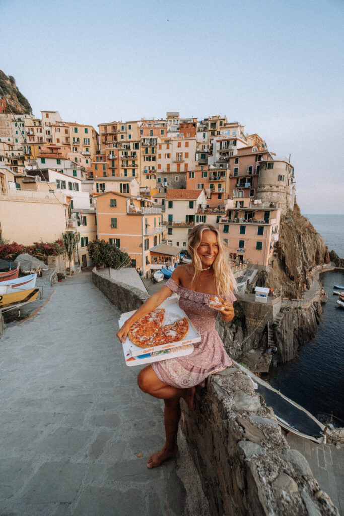Woman holding a pizza box and smiling on a rock wall of a pathway in Manarola. Behind is a view of Manarola village, with coloured buildings on a clifftop and harbour below.