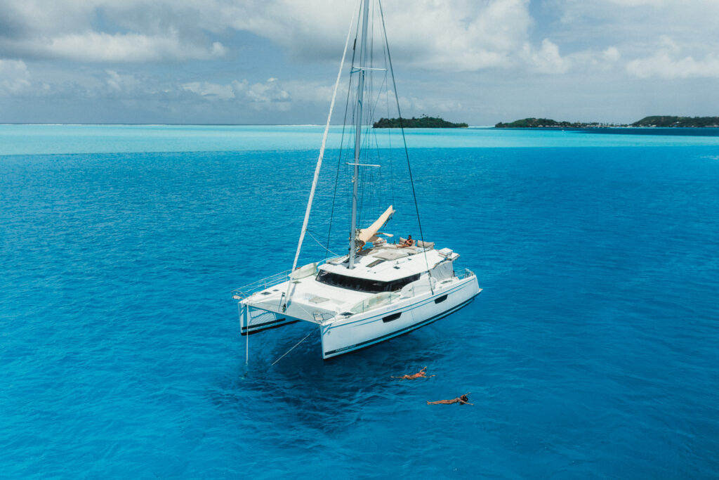 White catamaran boat on anchor inside the bright blue waters of the Bora Bora lagoon. Two woman float next to the boat in the water.