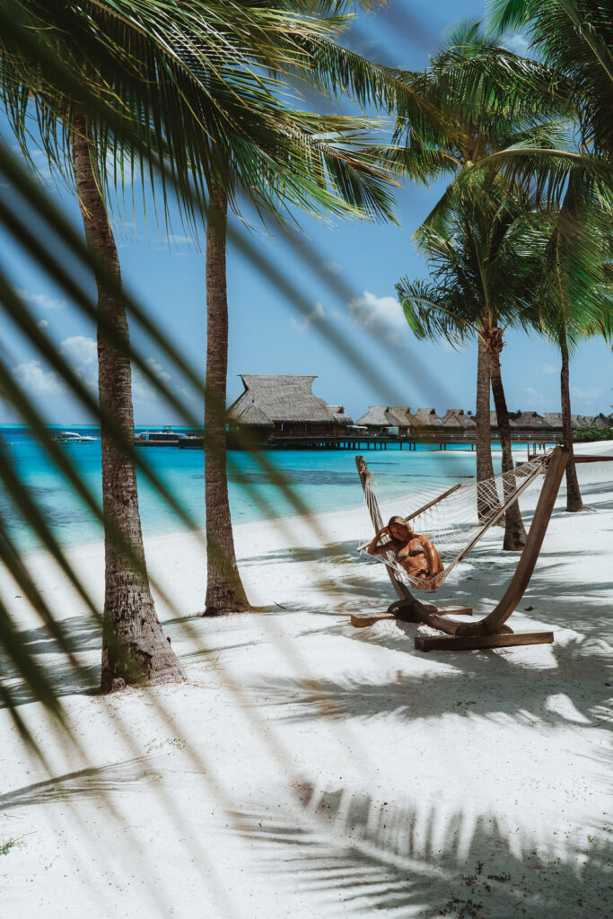 Woman sitting in a hammock beneath palm trees at a Bora Bora Resort on the beach. Calm turquoise water at the shore and villas in the distance