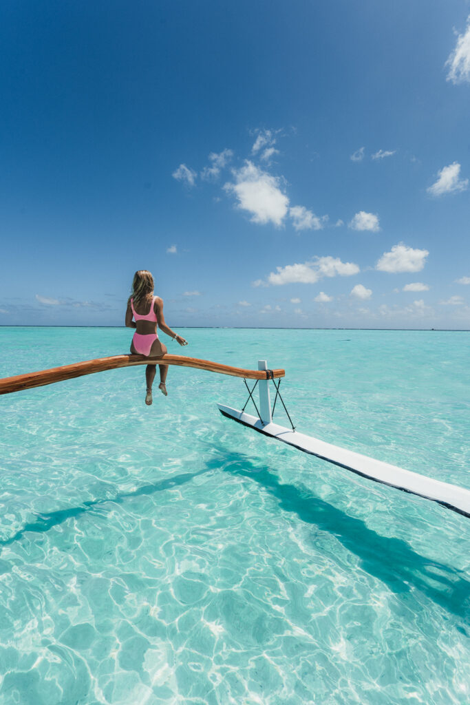 Woman sits on outrigger of tour boat with legs danggling over crystal clear lagoon.