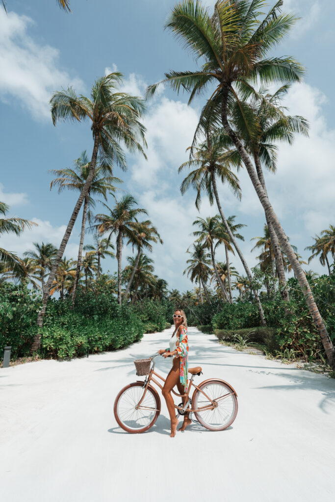 Woman standing on a bicycle on a white sand road that is lined with palm trees.