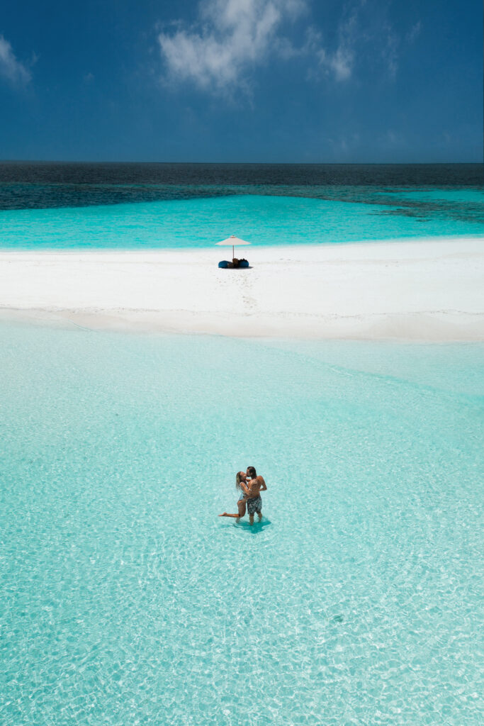 Couple kissing in shallow water, with a sandbank and private beach picnic set up behind them.