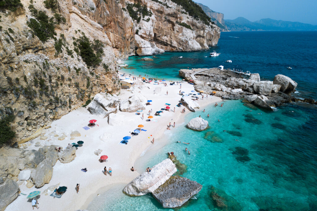 Aerial view of beach with colourful umbrellas, large boulders, and people swimming in clear blue waters.