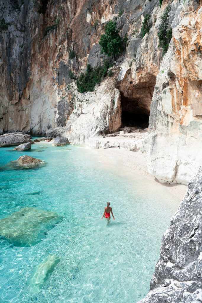 Woman wades through clear water, walking towards a cave.