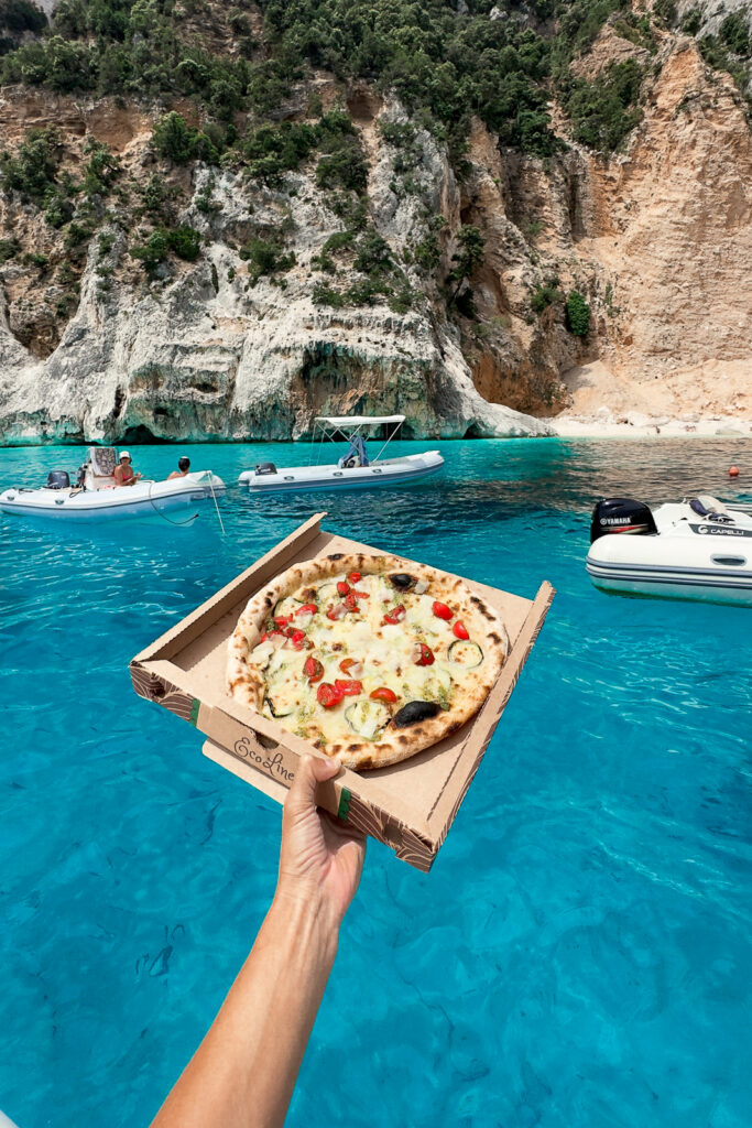 Woman's arm holding out a pizza in a takeaway box, extended over the water from her boat.