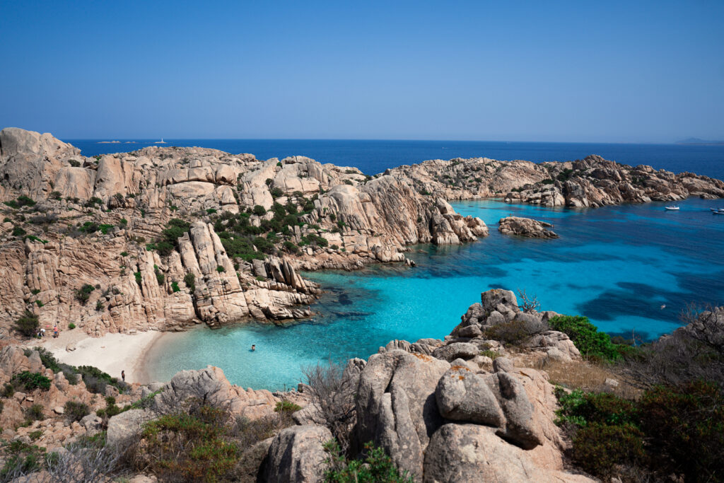 Cala Coticcio beach - a few bathers in blue water surrounded by rocky coastline.