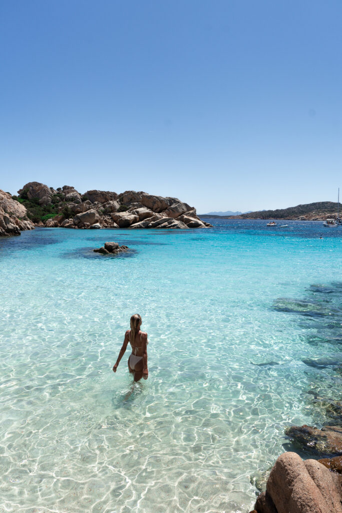 Cala Coticcio beach - woman enters the blue water surrounded by rocky coastline.