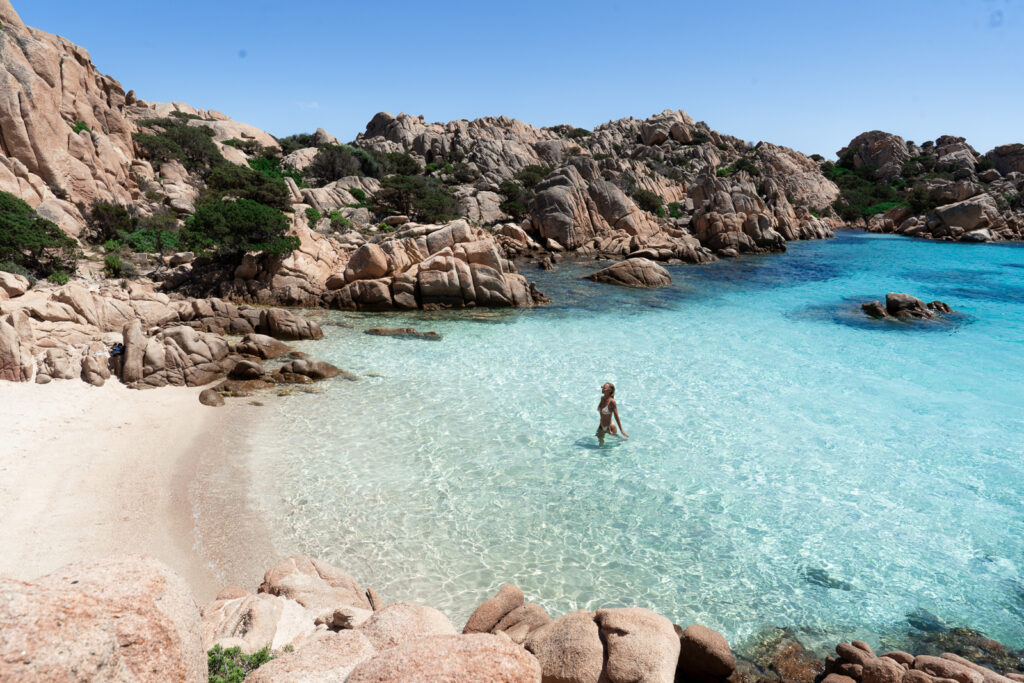 Cala Coticcio beach - a woman walking from the water in blue water surrounded by rocky coastline.