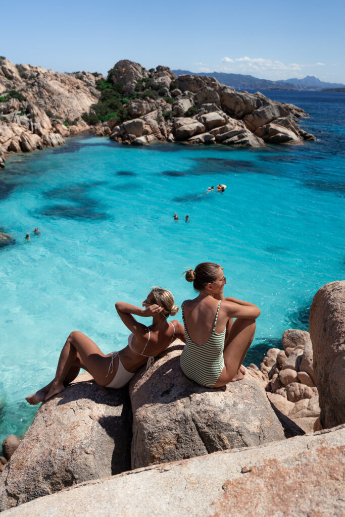 Two woman sitting on a rock look out over Cala Coticcio Beach below.