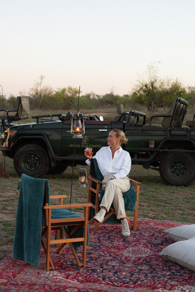 salty luxe enjoying a sunset drink sitting in a chair on a rug with pillows with a safari vehicle behind her in kruger national park