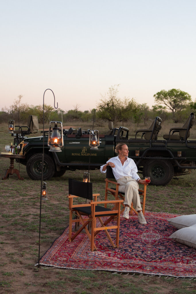 salty luxe sitting with a drink in hand and watching sunset with a safari vehicle from royal malewane behind her