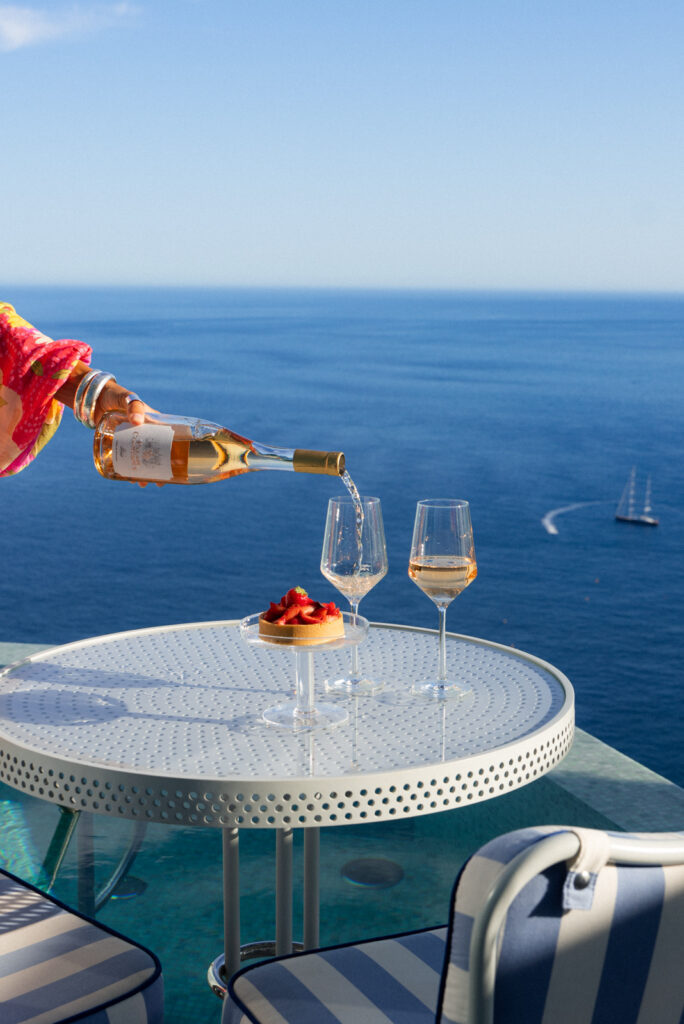 a person pours wine into a wine glass on a white table with the Mediterranean sea behind it at the maybourne riviera