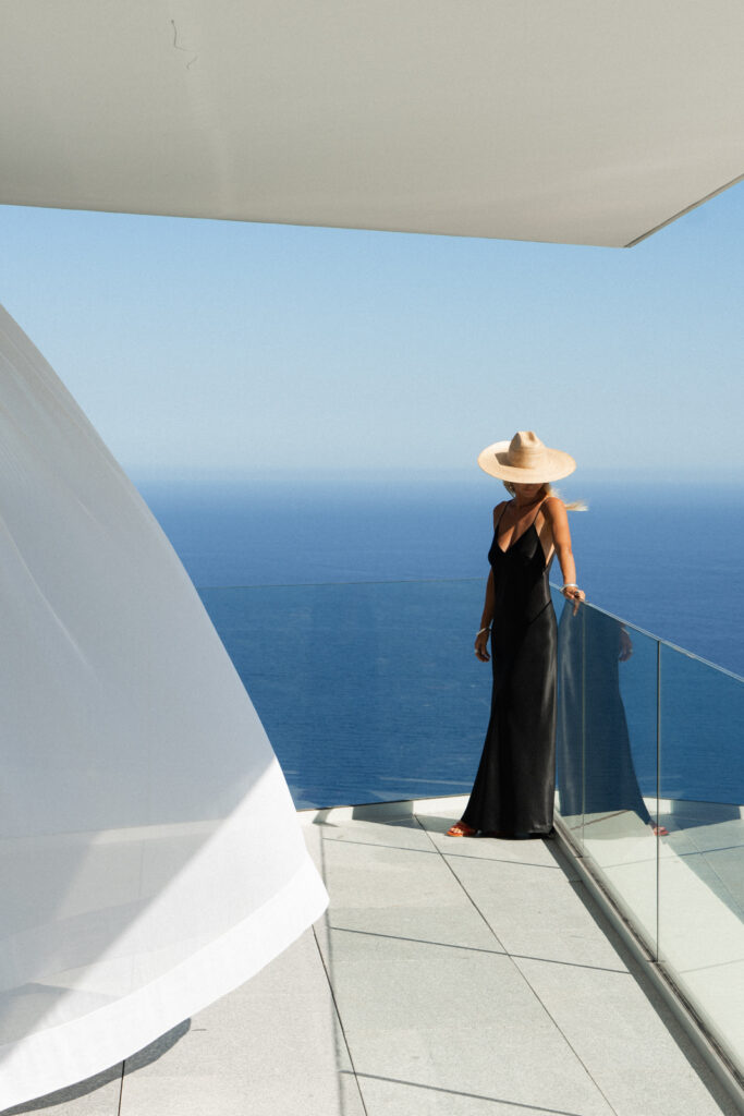 woman in a black dress and white hat standing at the edge of a balcony with the Mediterranean sea behind her at the maybourne riviera